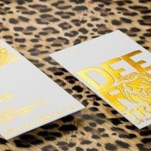 Puede incluir: Dos tarjetas de visita rectangulares blancas con letras en l&aacute;mina dorada y un gr&aacute;fico de le&oacute;n. El texto dice DEE ROCK TRAINING y DEREK CHRISTIE. Las tarjetas est&aacute;n colocadas sobre una superficie con estampado de leopardo marr&oacute;n y negro.