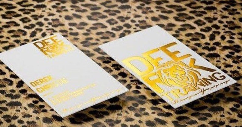 Puede incluir: Dos tarjetas de visita rectangulares blancas con letras en l&aacute;mina dorada y un gr&aacute;fico de le&oacute;n. El texto dice DEE ROCK TRAINING y DEREK CHRISTIE. Las tarjetas est&aacute;n colocadas sobre una superficie con estampado de leopardo marr&oacute;n y negro.