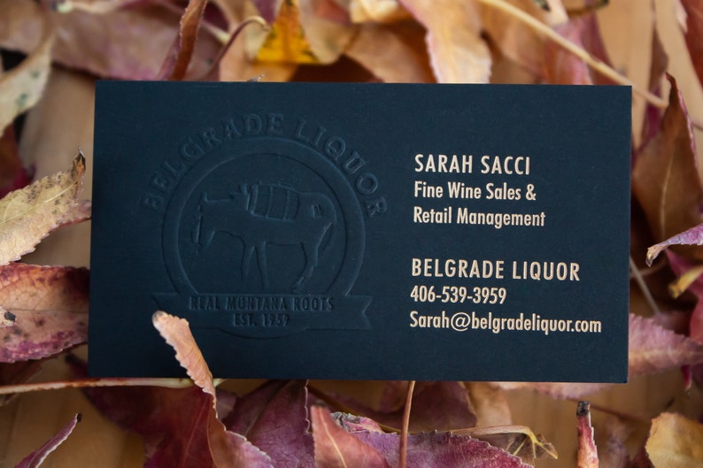 May include: A rectangular black business card with gold text reading Sarah Sacci, Fine Wine Sales & Retail Management, Belgrade Liquor, 406-539-3959, and Sarah@belgradeliquor.com. A debossed circular logo features a mule and the text Belgrade Liquor, Real Montana Roots, Est. 1957.