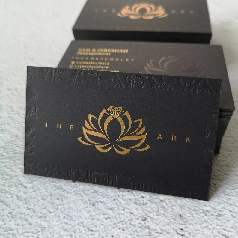 May include: A stack of rectangular black business cards featuring a gold foil logo of a lotus flower topped with a diamond. The text THE ARK is printed in gold. The border displays embossed Egyptian-style hieroglyphics and symbols.