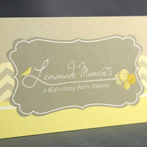 May include: A yellow and gray business card with a chevron pattern border. The card features a white, decorative frame with the text "Lemonade Moments, A Refreshing Party Paperie".