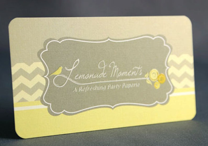 May include: A yellow and gray business card with a chevron pattern border. The card features a white, decorative frame with the text "Lemonade Moments, A Refreshing Party Paperie".