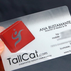 Może przedstawiać: Przezroczysta plastikowa wizyt&oacute;wka z czerwonym logo przedstawiającym stylizowaną figurę. Tekst na wizyt&oacute;wce brzmi "TallCat.com creativity-talent-community" i "ANA BUSTAMANTE Dyrektor ds. Medi&oacute;w i PR".