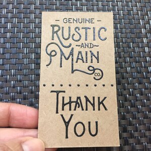 May include: A rectangular, tan-colored tag with black text. The top of the tag reads "GENUINE RUSTIC AND MAIN CO." in a decorative font. Below, the tag says "THANK YOU" in a bold, sans-serif font.