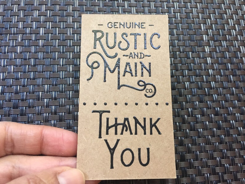 May include: A rectangular, tan-colored tag with black text. The top of the tag reads "GENUINE RUSTIC AND MAIN CO." in a decorative font. Below, the tag says "THANK YOU" in a bold, sans-serif font.