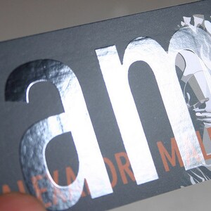 May include: A rectangular business card with a matte dark gray background. The letters am are printed in a large, reflective silver foil finish. Below, the text ALEXANDR MALPASS is printed in a copper-toned font, partially overlaid by a white graphic.