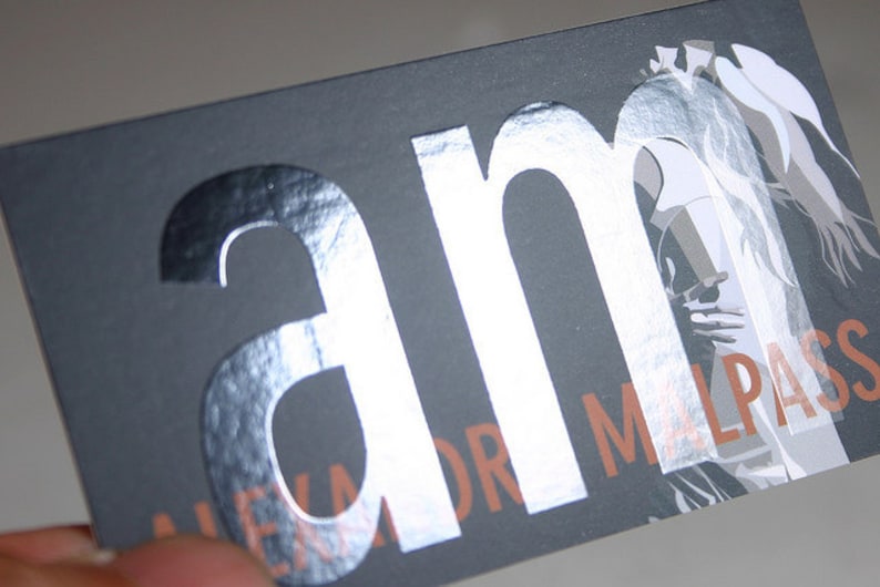 May include: A rectangular business card with a matte dark gray background. The letters am are printed in a large, reflective silver foil finish. Below, the text ALEXANDR MALPASS is printed in a copper-toned font, partially overlaid by a white graphic.
