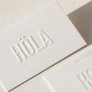 May include: A rectangular off-white business card featuring the word HÖLA embossed in large capital letters in the center. A second partially visible card below displays the text HÖLA STYLING CO in smaller embossed capital letters.