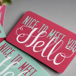 May include: A rectangular card with a dark pink background featuring the white script text Hello and the light blue sans-serif text Nice to meet you. The card has rounded corners and is displayed alongside other similar cards in light blue.