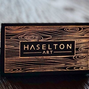May include: A stack of rectangular business cards featuring a black background with a metallic copper-colored wood grain pattern. The center of each card displays the text HASELTON ART in a clean, sans-serif font enclosed in a rectangular frame.