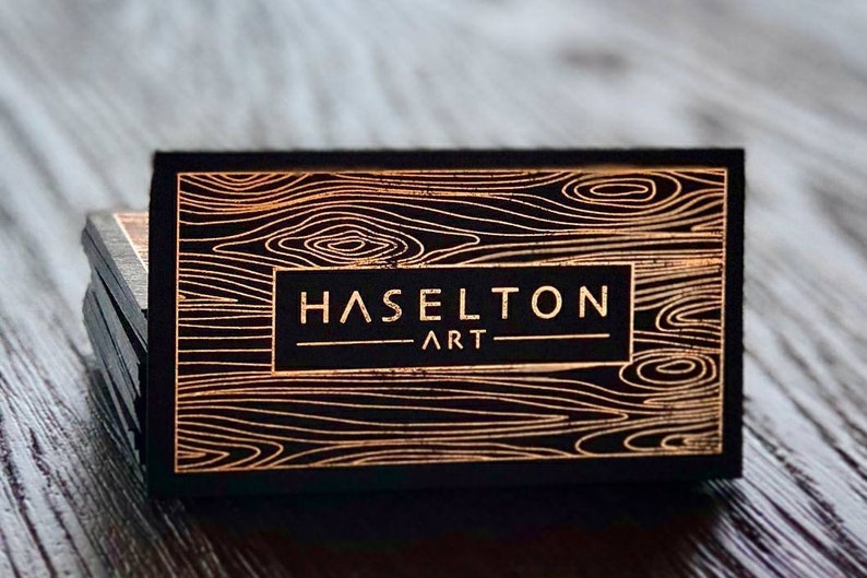 May include: A stack of rectangular business cards featuring a black background with a metallic copper-colored wood grain pattern. The center of each card displays the text HASELTON ART in a clean, sans-serif font enclosed in a rectangular frame.