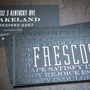 May include: Black business cards featuring the word FRESCOS in large white letters surrounded by a repeating pattern of the words ESCAPE, SATISFY, INDULGE, and ENJOY. One card displays the address 132 S KENTUCKY AVE, LAKELAND, and phone number 863-683-5267.