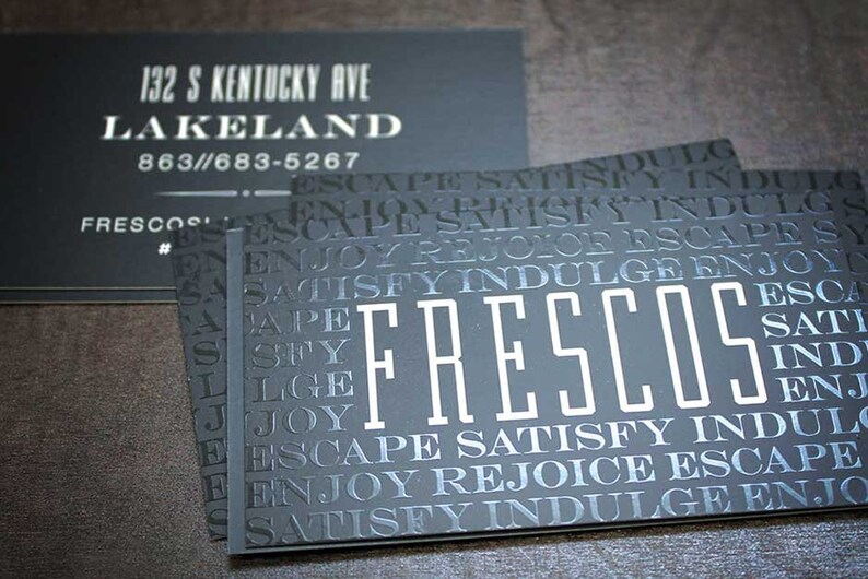May include: Black business cards featuring the word FRESCOS in large white letters surrounded by a repeating pattern of the words ESCAPE, SATISFY, INDULGE, and ENJOY. One card displays the address 132 S KENTUCKY AVE, LAKELAND, and phone number 863-683-5267.