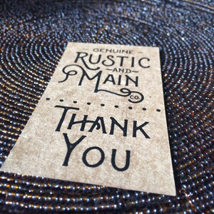May include: A brown paper tag with the words "GENUINE RUSTIC AND MAIN co." and "THANK YOU" in black lettering. The tag is on a dark, textured surface with a circular pattern of small, reflective beads.