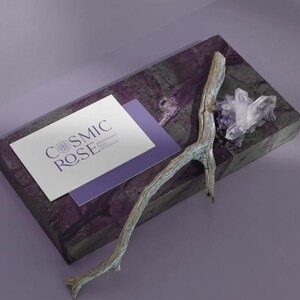 May include: A rectangular purple stone slab featuring a white business card with the text COSMIC ROSE, a weathered grey wooden twig, and a cluster of clear quartz crystals. The card also displays smaller text reading BREATHWORK REIKI MEDITATION.