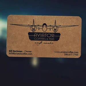 May include: A rectangular business card with rounded corners. The card is a light brown color and features a black graphic of a vintage airplane above the words "AVIATOR COFFEES & TEAS craft roaster". The owner's name, email, and website are also printed on the card.
