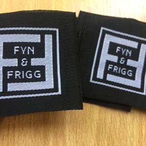 May include: Two square black fabric labels featuring a white embroidered logo. The design consists of a stylized letter F inside a square border, with the text FYN & FRIGG centered within the letter. The embroidery is clean and clearly defined.