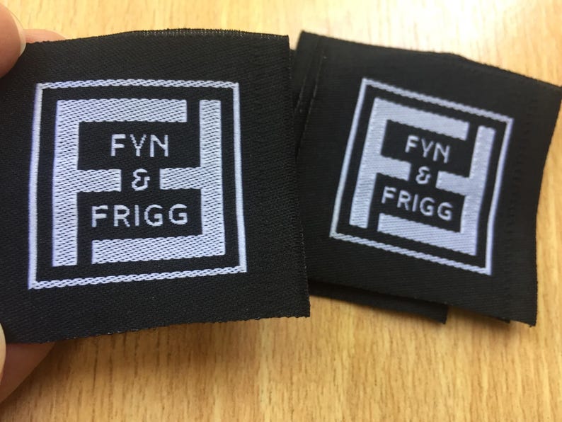 May include: Two square black fabric labels featuring a white embroidered logo. The design consists of a stylized letter F inside a square border, with the text FYN & FRIGG centered within the letter. The embroidery is clean and clearly defined.