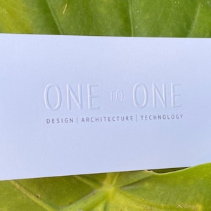 Könnte beinhalten: Eine hellblaue Visitenkarte mit dem geprägten Text "ONE to ONE" über den Worten "DESIGN | ARCHITECTURE | TECHNOLOGY". Die Karte liegt vor einem grünen Blatt-Hintergrund.