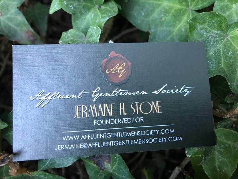K&ouml;nnte beinhalten: Eine rechteckige schwarze Visitenkarte mit einem roten Wachssiegel-Motiv und goldenen Initialen AG oben. Der Text lautet Affluent Gentlemen Society, Jermaine H. Stone, Founder/Editor, inklusive Website und E-Mail-Adresse in wei&szlig;er Schrift.
