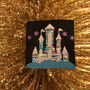 May include: A square black fabric patch featuring an embroidered castle design. The castle has towers with white roofs and a multi-colored, iridescent, metallic-thread body. A solid teal blue base sits below the castle, and small purple embroidered stars are positioned on either side of the central tower against the black background.