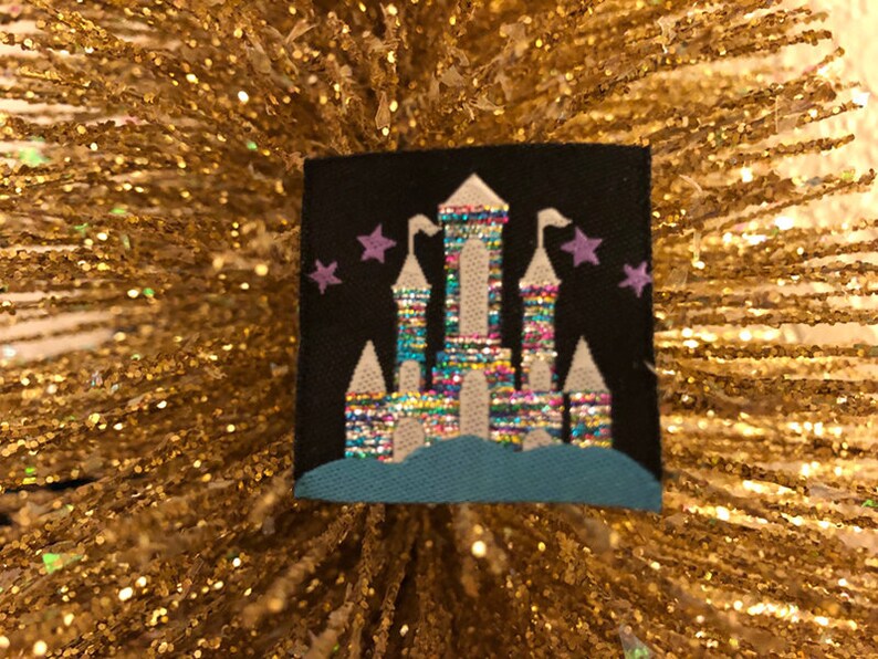 May include: A square black fabric patch featuring an embroidered castle design. The castle has towers with white roofs and a multi-colored, iridescent, metallic-thread body. A solid teal blue base sits below the castle, and small purple embroidered stars are positioned on either side of the central tower against the black background.