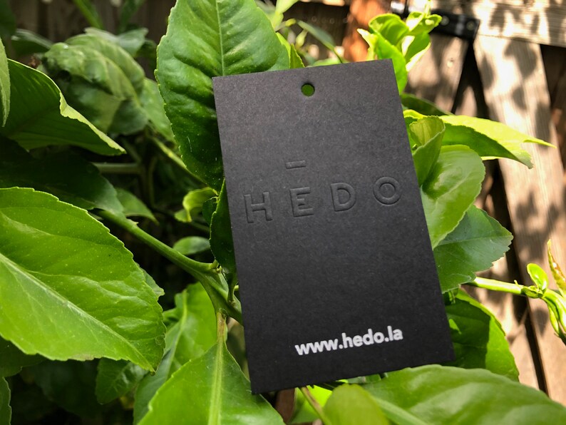 May include: A rectangular black paper tag featuring a small circular hole at the top. The word HEDO is embossed in the center with a horizontal line above the letter E. The website address www.hedo.la is printed in white at the bottom of the tag.