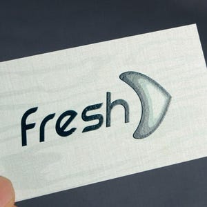 K&ouml;nnte beinhalten: Eine rechteckige wei&szlig;e Karte mit dem Wort fresh in dunkelblauen Kleinbuchstaben, gefolgt von einem stilisierten grauen Symbol in Form eines Bumerangs. Die Karte weist eine subtile horizontale Holzmaserung auf der gesamten Oberfl&auml;che auf.