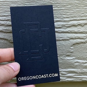 May include: A rectangular black business card featuring a large, embossed, tonal monogram logo in the center. The bottom of the card displays the white text OREGONCOAST.COM in a clean, sans-serif font, providing a clear and professional look.