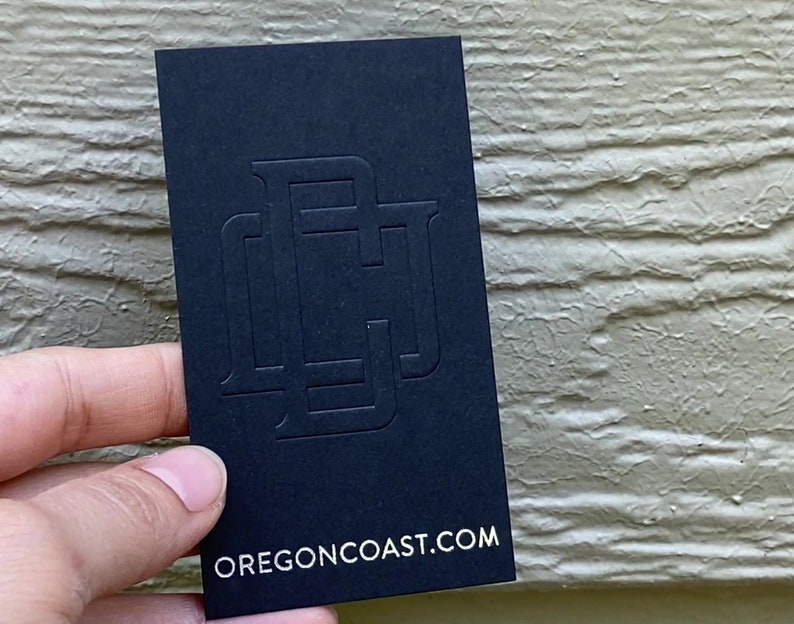 May include: A rectangular black business card featuring a large, embossed, tonal monogram logo in the center. The bottom of the card displays the white text OREGONCOAST.COM in a clean, sans-serif font, providing a clear and professional look.