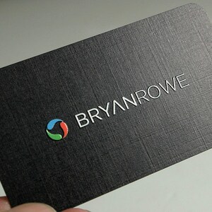 K&ouml;nnte beinhalten: Eine rechteckige schwarze Visitenkarte mit strukturierter Oberfl&auml;che. Die Karte zeigt den wei&szlig;en Schriftzug BRYANROWE und ein kreisf&ouml;rmiges Logo aus drei geschwungenen Segmenten in Blau, Rot und Gr&uuml;n. Die Ecken der Karte sind abgerundet.