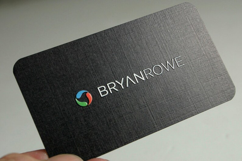 K&ouml;nnte beinhalten: Eine rechteckige schwarze Visitenkarte mit strukturierter Oberfl&auml;che. Die Karte zeigt den wei&szlig;en Schriftzug BRYANROWE und ein kreisf&ouml;rmiges Logo aus drei geschwungenen Segmenten in Blau, Rot und Gr&uuml;n. Die Ecken der Karte sind abgerundet.