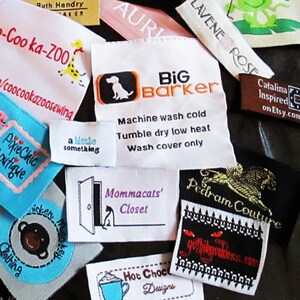 May include: A collection of various rectangular fabric clothing labels featuring diverse brand names and logos including Big Barker, Coo-ka-ZOO, PixieChic Boutique, Mommacats' Closet, Pedram Couture, and Hot Choco Designs with embroidered text and graphics.