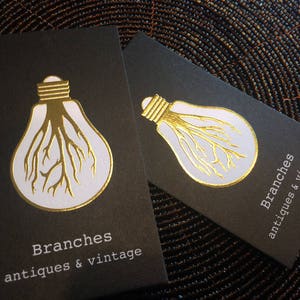 Puede incluir: Dos tarjetas de visita rectangulares negras con un gr&aacute;fico en l&aacute;mina dorada de una bombilla con ra&iacute;ces de &aacute;rbol en su interior. El texto Branches antiques & vintage est&aacute; impreso en blanco en la parte inferior de cada tarjeta, con un dise&ntilde;o limpio.
