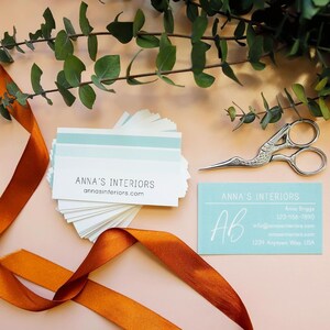 May include: A stack of white business cards with a light blue stripe and the text ANNA'S INTERIORS and annasinteriors.com, alongside a single light blue card with white text, a silver crane-shaped pair of scissors, and a burnt orange ribbon.