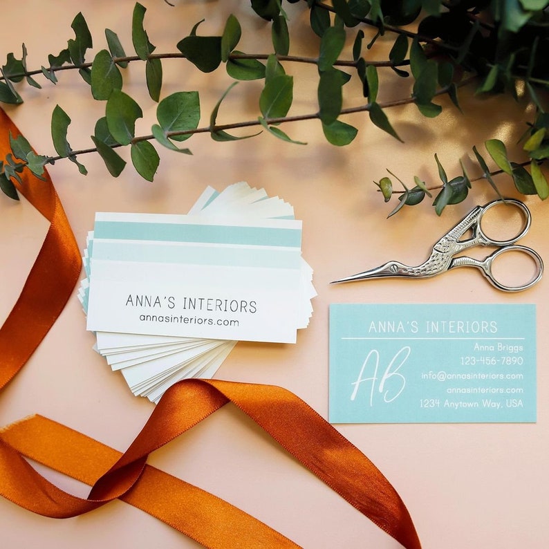 May include: A stack of white business cards with a light blue stripe and the text ANNA'S INTERIORS and annasinteriors.com, alongside a single light blue card with white text, a silver crane-shaped pair of scissors, and a burnt orange ribbon.