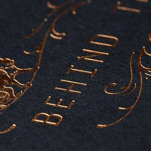 May include: A dark charcoal textured paper surface featuring metallic copper foil stamping. The design includes a stylized illustration of a small animal and elegant swirling flourishes alongside the partial text BEHIND T, rendered in a clean serif font.