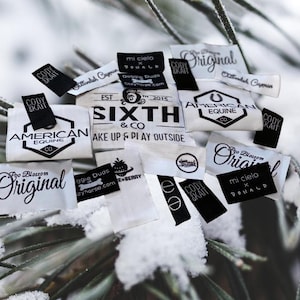 May include: A collection of small, rectangular fabric clothing labels in black and white. The labels feature various printed brand names and logos, including Sixth & Co, American Equine, and Poo Blossom Original, with some text reading Wake Up & Play Outside.