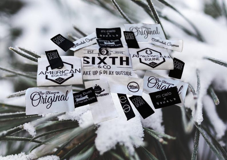 May include: A collection of small, rectangular fabric clothing labels in black and white. The labels feature various printed brand names and logos, including Sixth & Co, American Equine, and Poo Blossom Original, with some text reading Wake Up & Play Outside.
