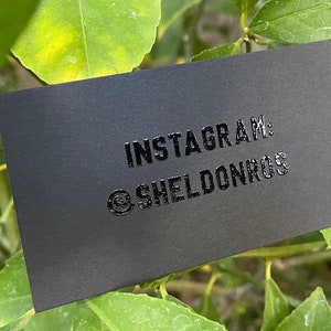 May include: A rectangular matte black business card featuring glossy raised lettering that reads INSTAGRAM: @SHELDONROS. The card is positioned against a backdrop of green leaves, with the text centered clearly on the dark surface of the card.