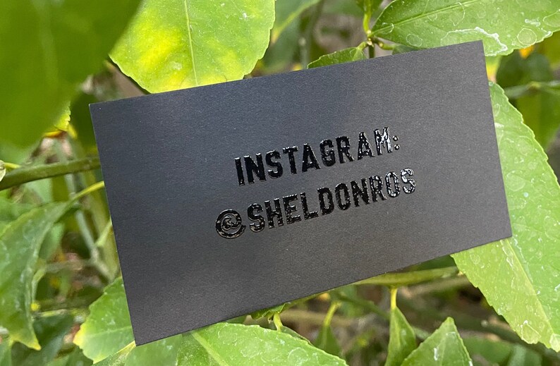 May include: A rectangular matte black business card featuring glossy raised lettering that reads INSTAGRAM: @SHELDONROS. The card is positioned against a backdrop of green leaves, with the text centered clearly on the dark surface of the card.