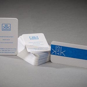 以下が含まれることがあります： 角が丸い白い名刺の束。K AND K GRAPHICS、photography、design、calligraphyの文字が入った青いロゴが特徴です。1枚は垂直に立ち、もう1枚は青い帯が入った状態で水平に置かれています。