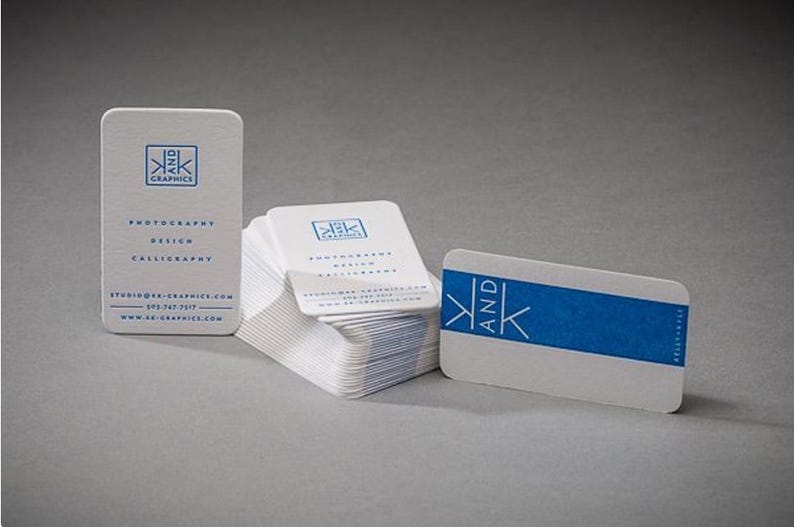 以下が含まれることがあります： 角が丸い白い名刺の束。K AND K GRAPHICS、photography、design、calligraphyの文字が入った青いロゴが特徴です。1枚は垂直に立ち、もう1枚は青い帯が入った状態で水平に置かれています。