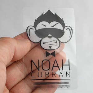Puede incluir: Una tarjeta rectangular de pl&aacute;stico transparente con un gr&aacute;fico negro de un mono con gafas de sol y pajarita. Debajo del mono, el texto NOAH CURRAN est&aacute; impreso en una fuente negra sans-serif clara, con una l&iacute;nea horizontal debajo.