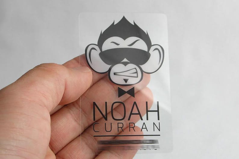Puede incluir: Una tarjeta rectangular de pl&aacute;stico transparente con un gr&aacute;fico negro de un mono con gafas de sol y pajarita. Debajo del mono, el texto NOAH CURRAN est&aacute; impreso en una fuente negra sans-serif clara, con una l&iacute;nea horizontal debajo.