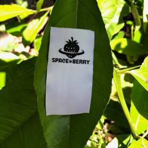 May include: A rectangular white fabric clothing label featuring a black graphic of a strawberry shaped like a flying saucer. Below the graphic, the text reads SPACE BERRY in a bold, sans-serif font. The label is positioned on a green leaf.