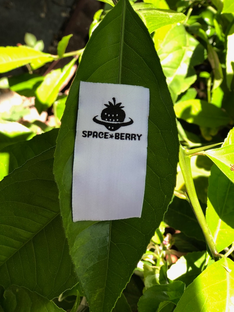 May include: A rectangular white fabric clothing label featuring a black graphic of a strawberry shaped like a flying saucer. Below the graphic, the text reads SPACE BERRY in a bold, sans-serif font. The label is positioned on a green leaf.