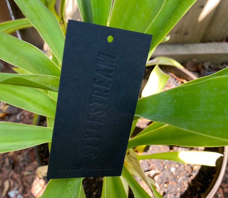 May include: A rectangular black hang tag featuring the embossed text STYLESTREAM in a vertical orientation. The tag includes a small circular hole at the top for attachment and the website URL WWW.SHOPSTYLESTREAM.COM printed at the bottom.