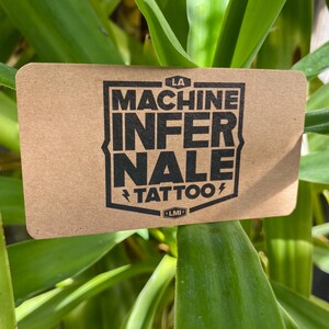 May include: A rectangular tan card with rounded corners featuring a black shield-shaped logo. The text reads LA MACHINE INFERNALE TATTOO with small lightning bolt icons on either side of the word TATTOO and a small LMI emblem at the bottom.