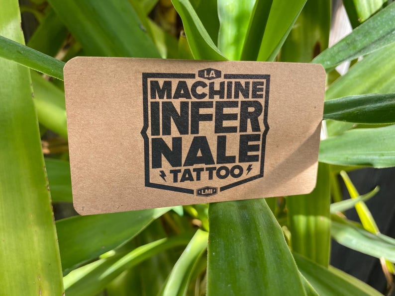 May include: A rectangular tan card with rounded corners featuring a black shield-shaped logo. The text reads LA MACHINE INFERNALE TATTOO with small lightning bolt icons on either side of the word TATTOO and a small LMI emblem at the bottom.